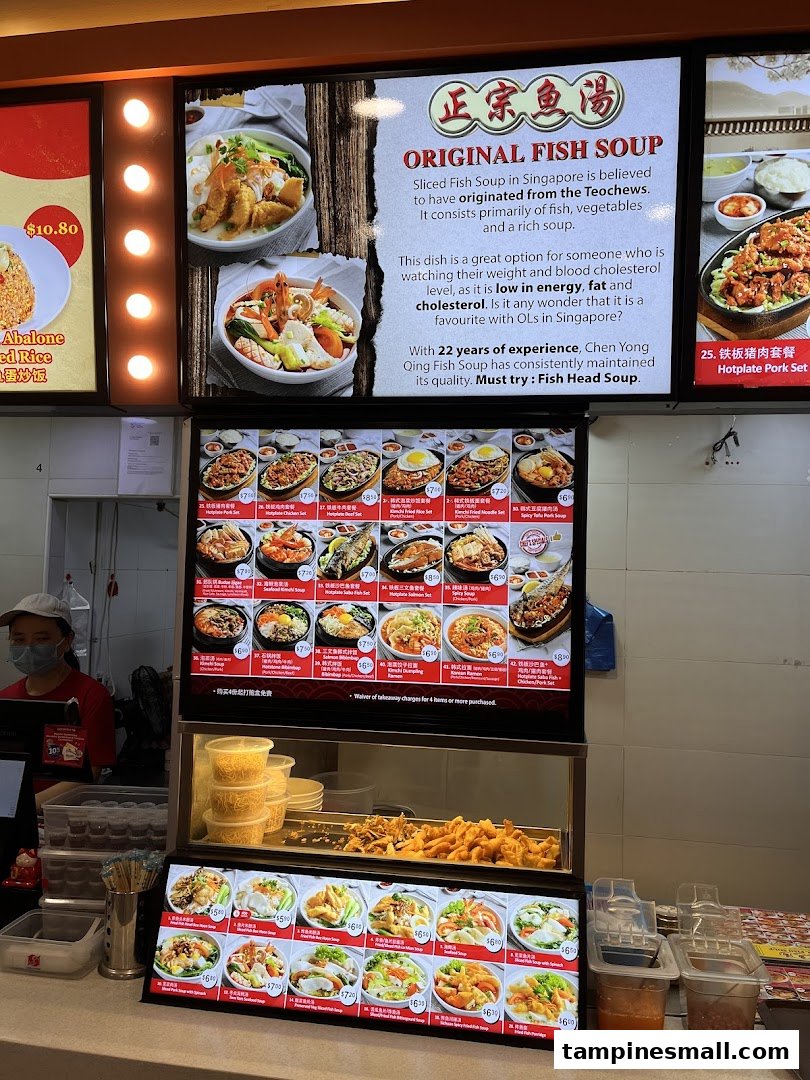 A food stall counter with digital menu boards displaying various fish soup dishes and a staff member.
