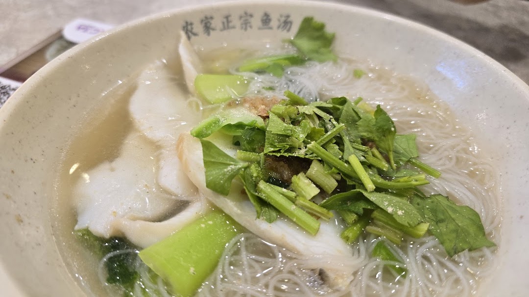 A close-up of a bowl of fish soup with thin noodles, fish slices, and green vegetables.