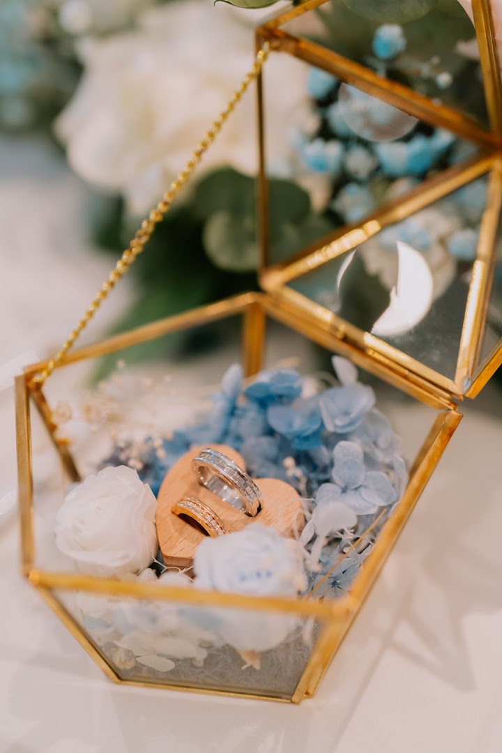 A close-up of two wedding rings displayed in a geometric glass box with blue and white flowers.