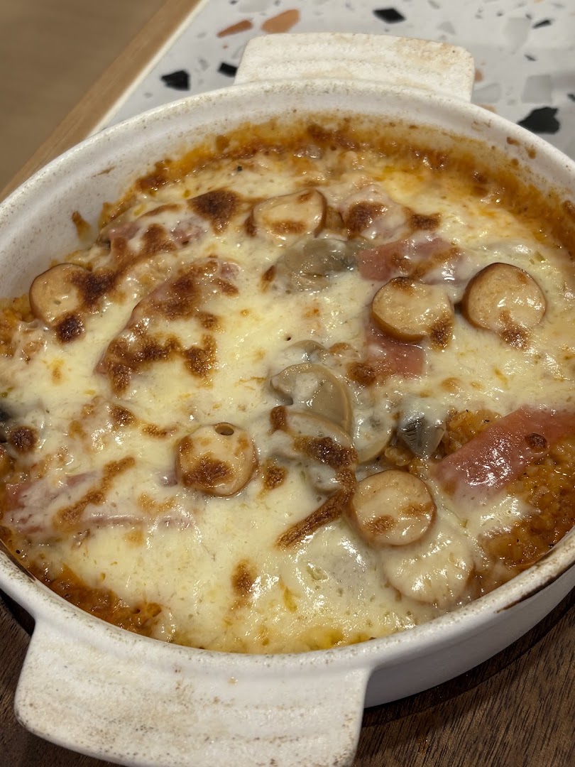 A close-up of a cheesy baked dish with sausages and mushrooms in a white ceramic bowl.