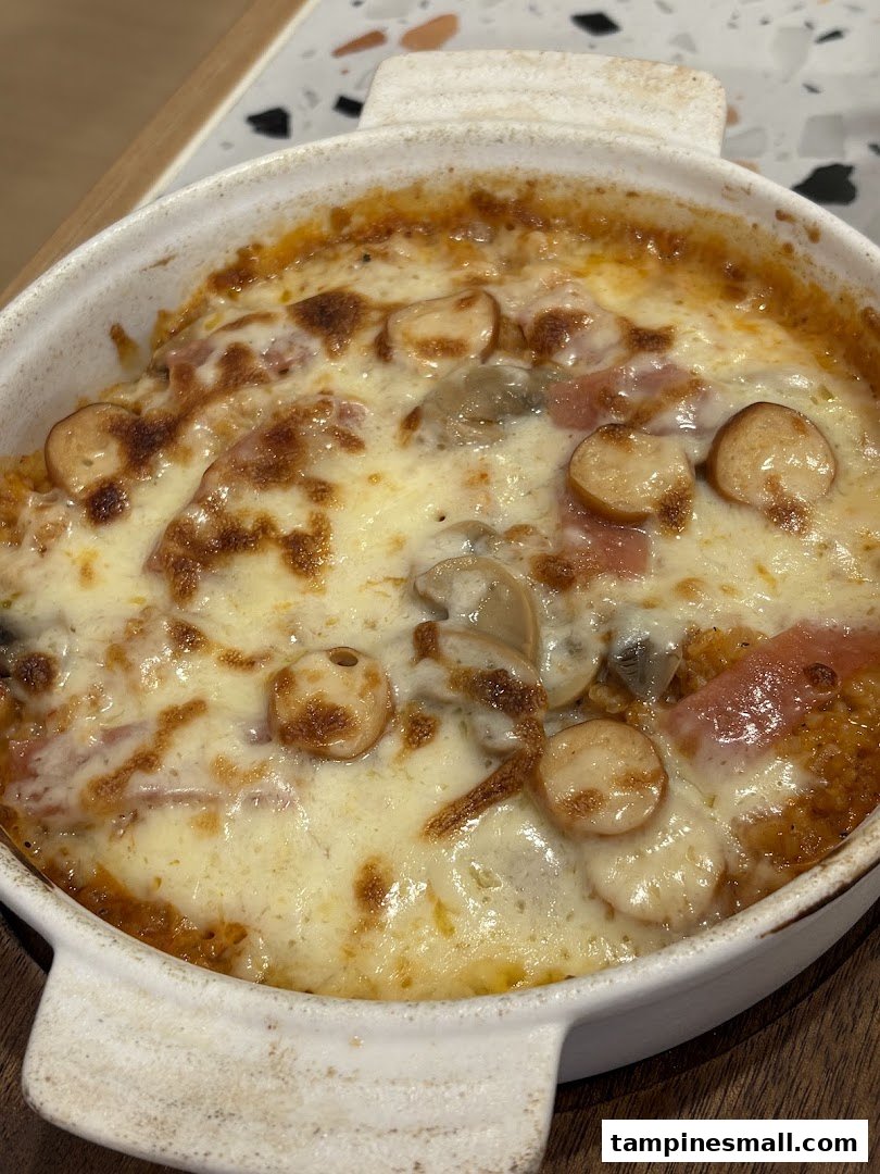 A close-up of a cheesy baked dish with sausages and mushrooms in a white ceramic bowl.