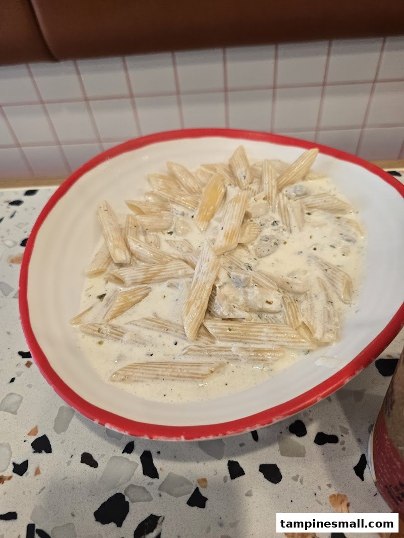 A close-up of penne pasta in a creamy white sauce, served in a white bowl with a red rim.