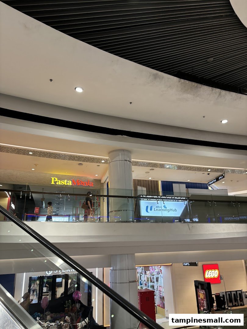 The PastaMania shopfront is visible on an upper level of a bustling shopping mall, viewed from an escalator.