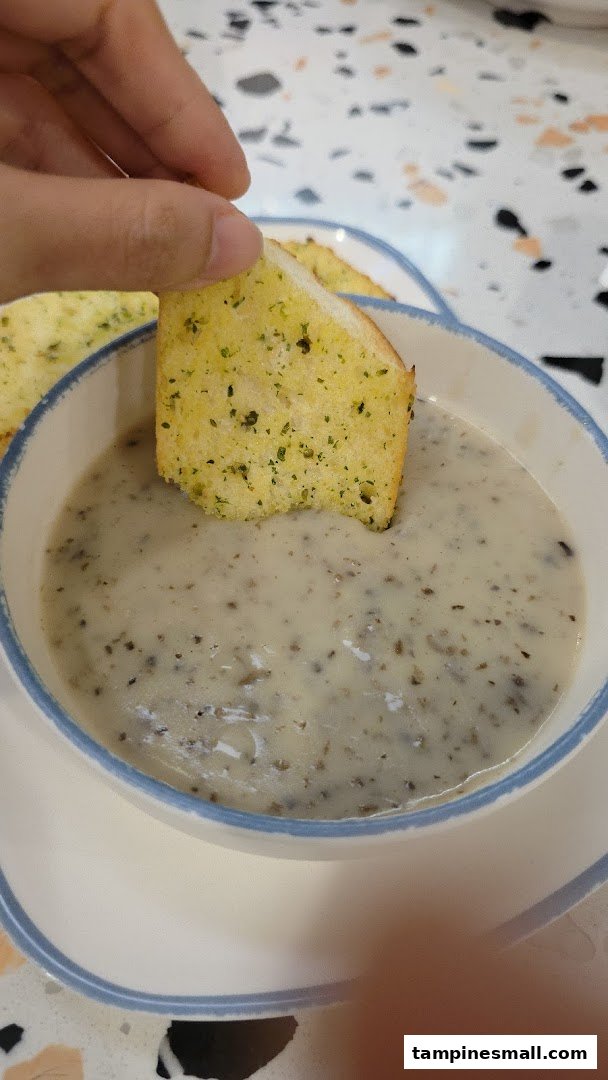 A hand dips a piece of garlic bread into a creamy bowl of mushroom soup.