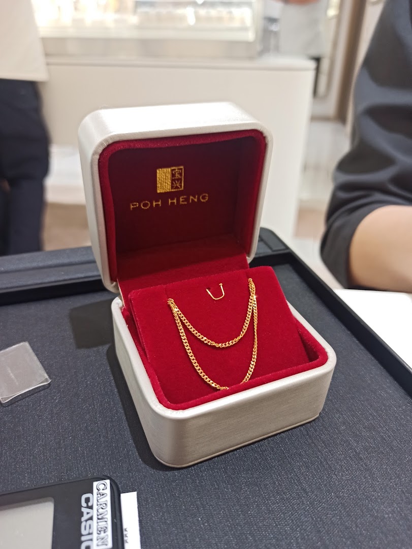 A delicate gold chain displayed in a red velvet Poh Heng jewellery box on a dark counter.
