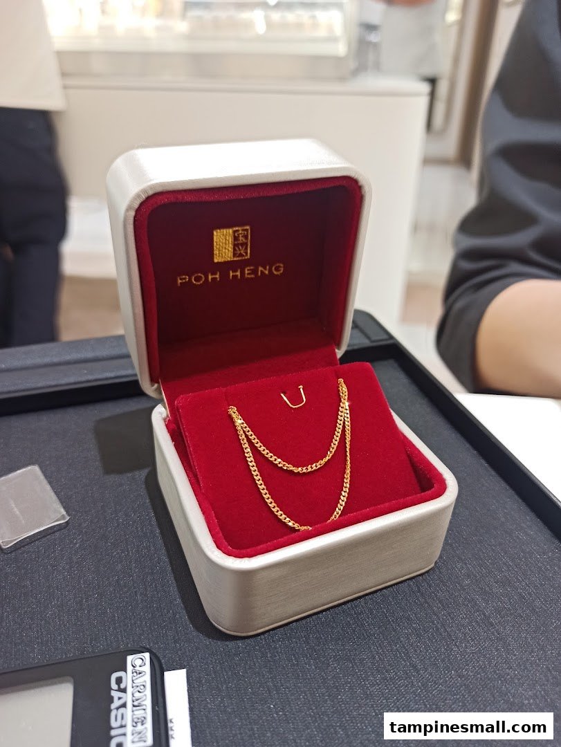 A delicate gold chain displayed in a red velvet Poh Heng jewellery box on a dark counter.