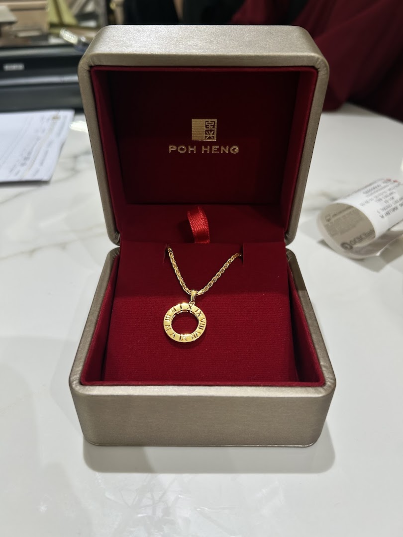 A gold necklace with a Roman numeral pendant in a red and gold Poh Heng box.