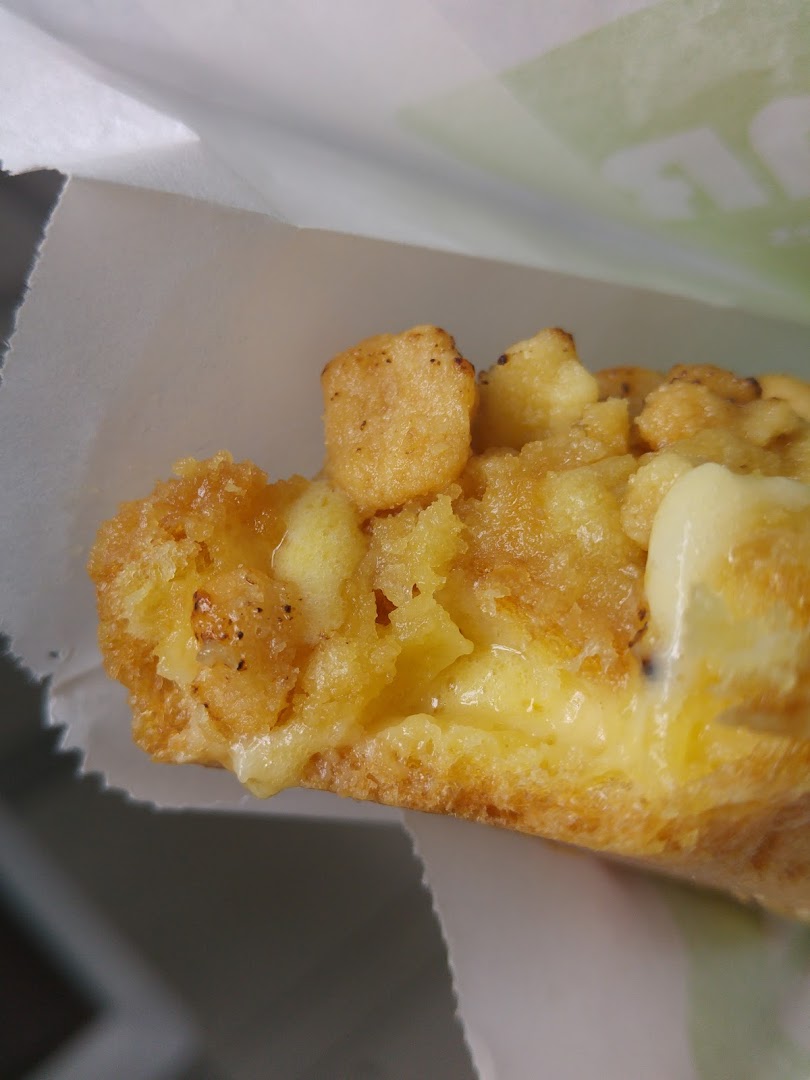 A close-up shot of a golden-brown pastry with a creamy filling and crispy topping, served in a white paper bag.