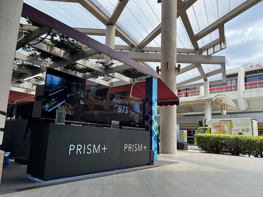 An outdoor display booth for PRISM+ featuring large screens, located under a covered walkway near a train track.
