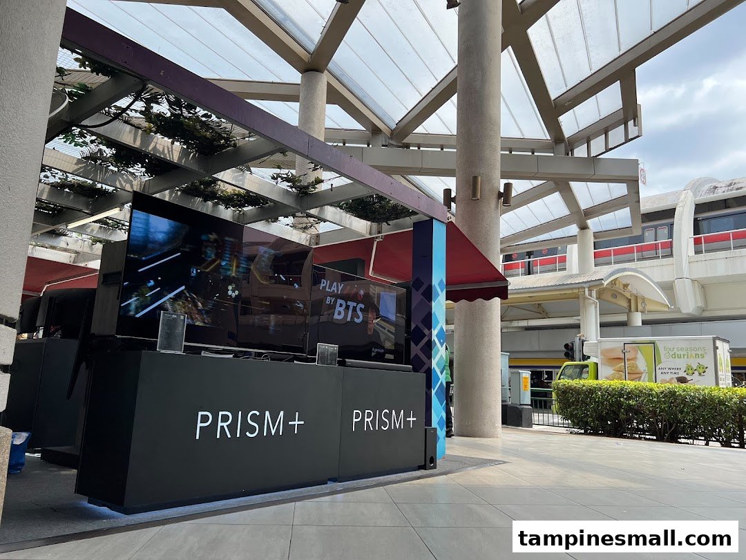 An outdoor display booth for PRISM+ featuring large screens, located under a covered walkway near a train track.