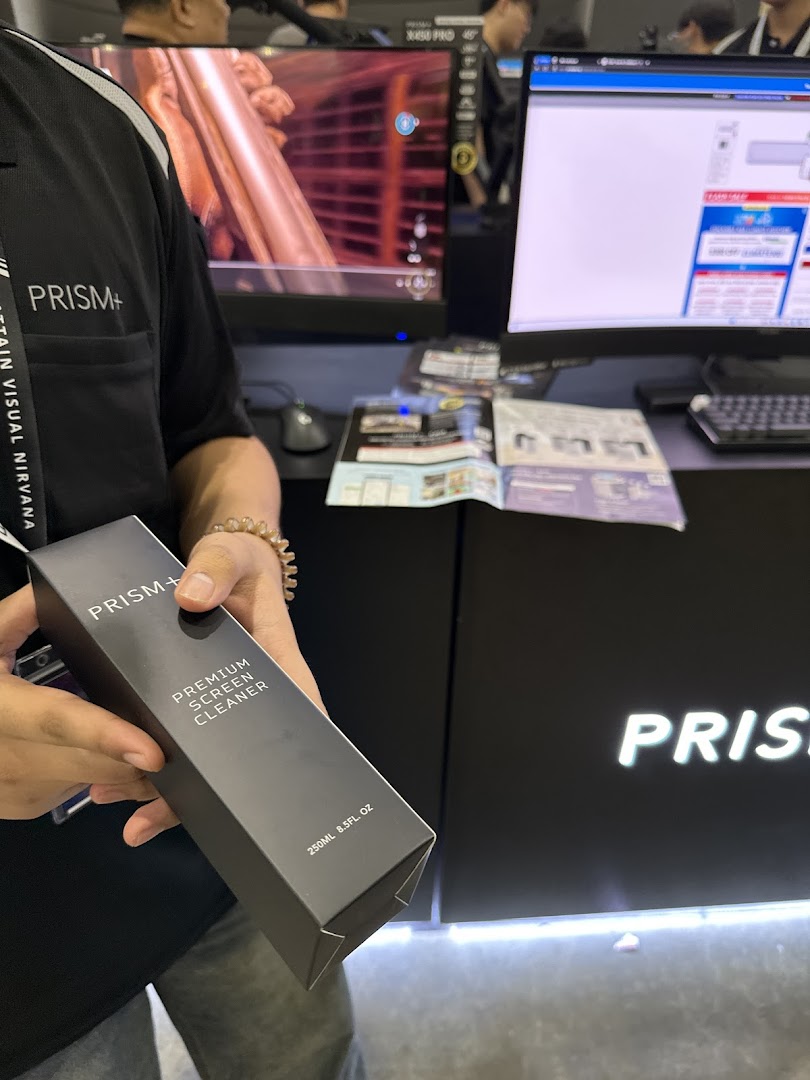 A person holds a PRISM+ Premium Screen Cleaner in a tech shop featuring monitors and the PRISM+ logo.