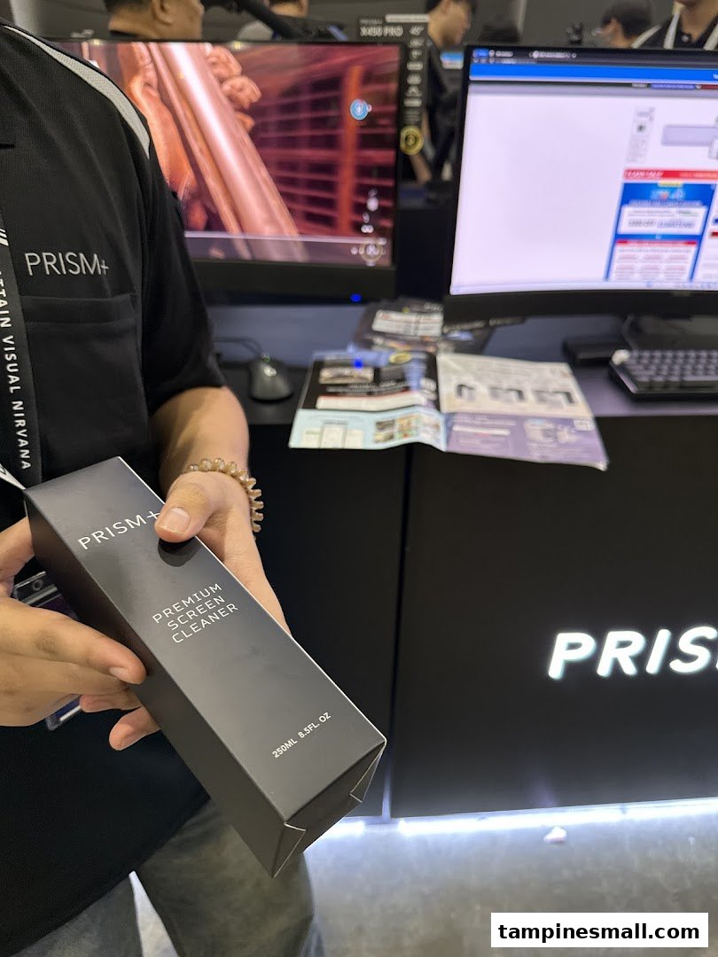 A person holds a PRISM+ Premium Screen Cleaner in a tech shop featuring monitors and the PRISM+ logo.