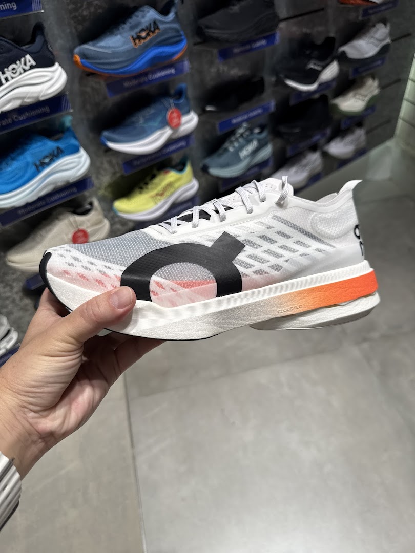 A hand holds a white running shoe with orange accents, showcasing footwear inside Running Lab.