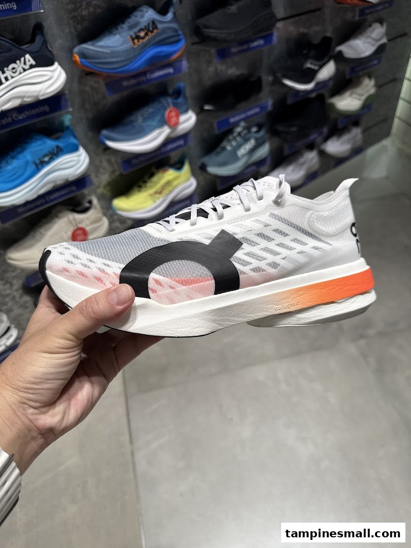 A hand holds a white running shoe with orange accents, showcasing footwear inside Running Lab.