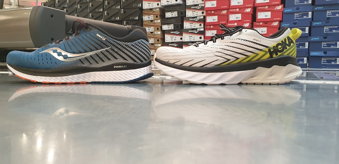 Two running shoes, a blue Saucony and a white Hoka, displayed on a reflective floor inside a shoe store.