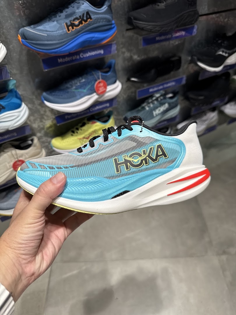 A hand holds a light blue Hoka running shoe inside Running Lab, with other shoes displayed.