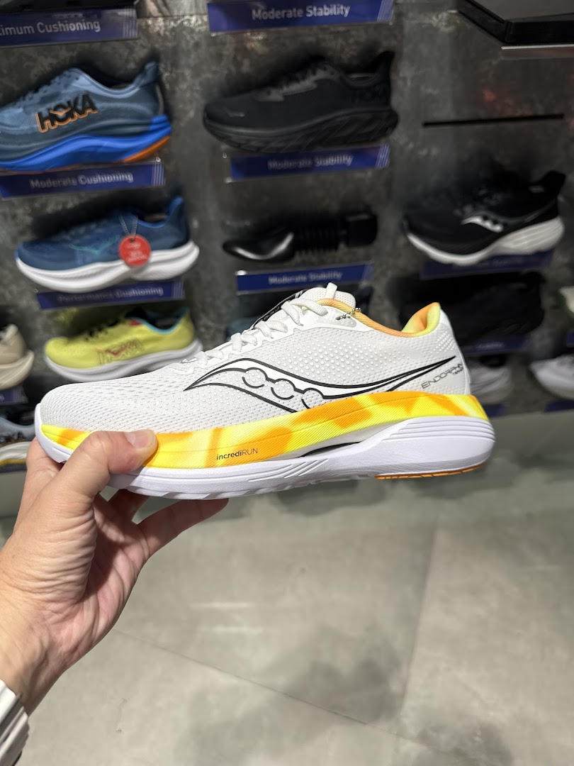 A hand holds a white running shoe with a vibrant yellow sole, displayed against a wall of athletic footwear.