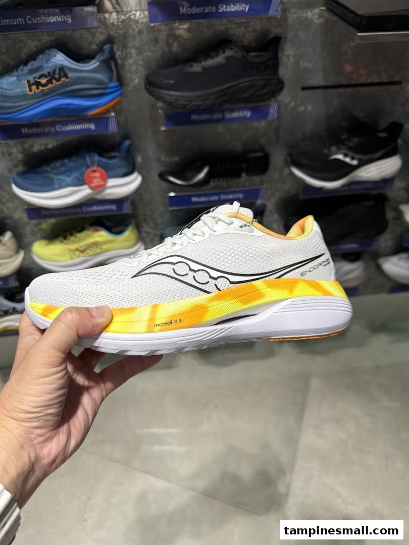 A hand holds a white running shoe with a vibrant yellow sole, displayed against a wall of athletic footwear.