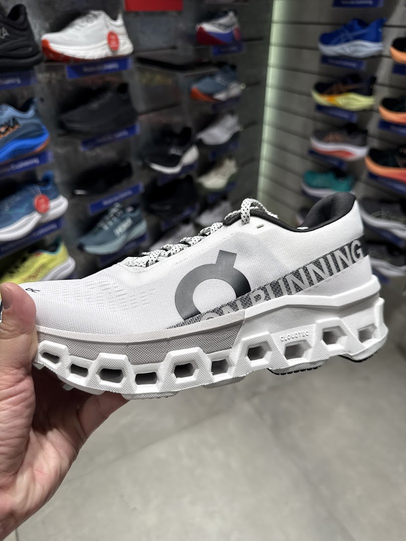 A hand holds a white running shoe with a unique sole, showcasing other shoes in the background.