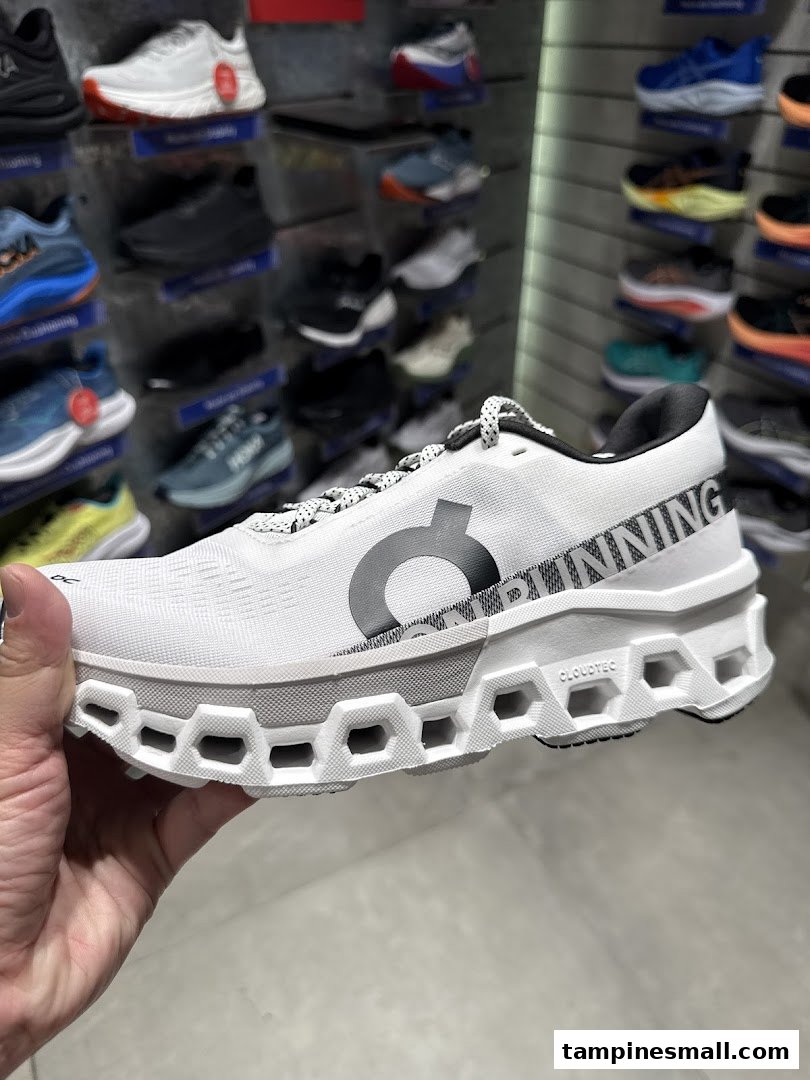 A hand holds a white running shoe with a unique sole, showcasing other shoes in the background.