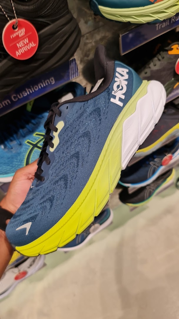 A hand holds a blue and neon green Hoka running shoe inside Running Lab, showcasing new arrivals.