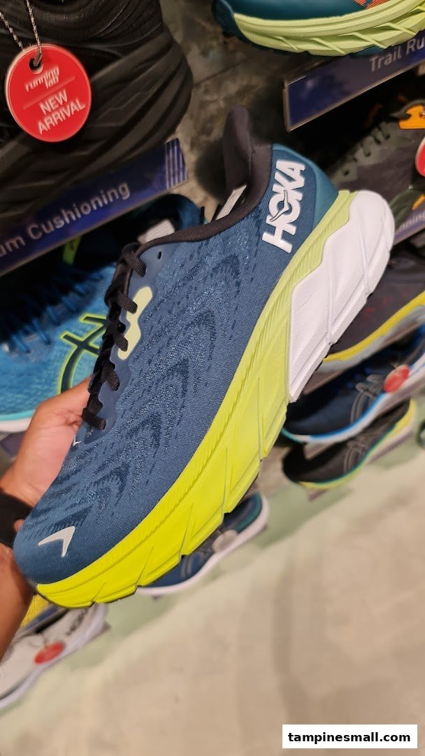 A hand holds a blue and neon green Hoka running shoe inside Running Lab, showcasing new arrivals.