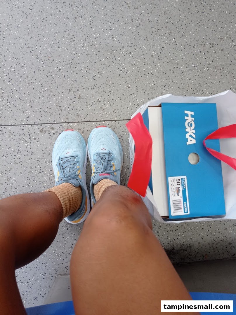 A person wearing new light blue Hoka running shoes with the shoe box and shopping bag nearby.