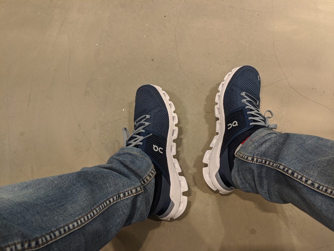 A person's feet wearing blue On running shoes with white soles, likely trying them on.