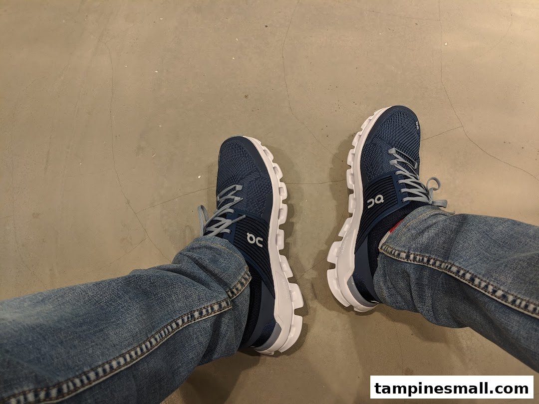 A person's feet wearing blue On running shoes with white soles, likely trying them on.