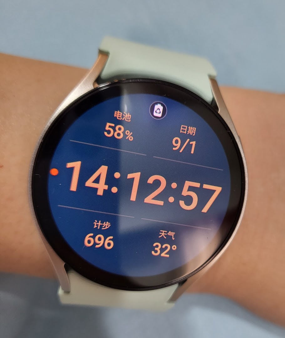 A close-up of a Samsung smartwatch on a wrist, displaying time, battery, date, steps, and weather.