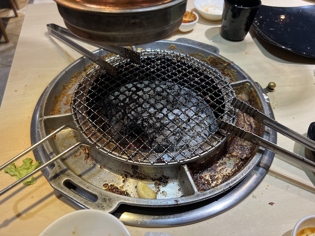 A close-up of a charcoal grill with tongs and side dishes on a table, ready for Korean BBQ.