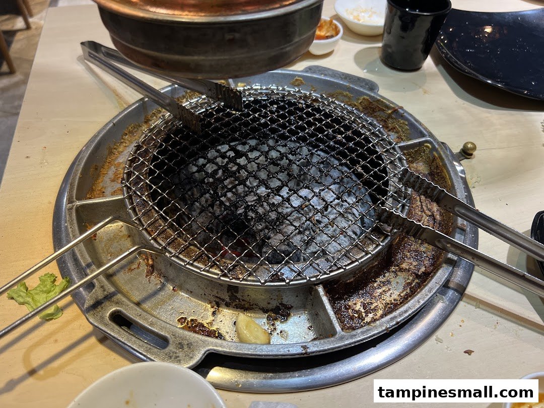 A close-up of a charcoal grill with tongs and side dishes on a table, ready for Korean BBQ.