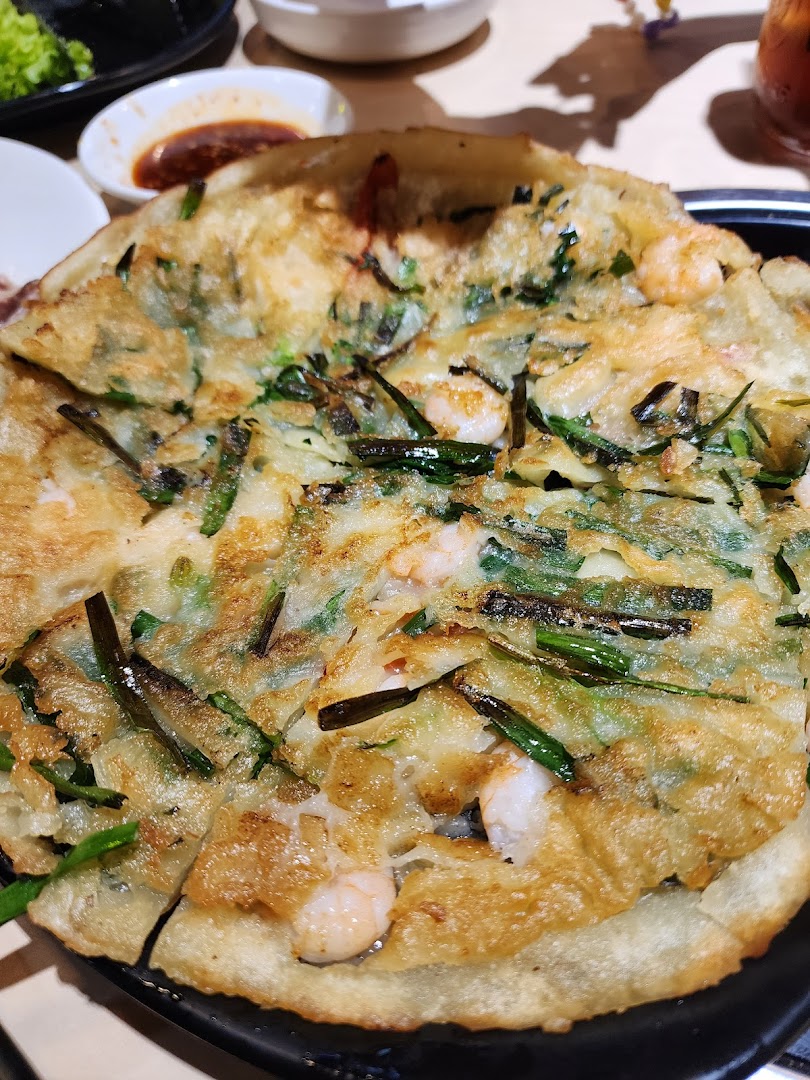 A close-up of a golden-brown Korean seafood and green onion pancake (haemul pajeon) on a black plate.