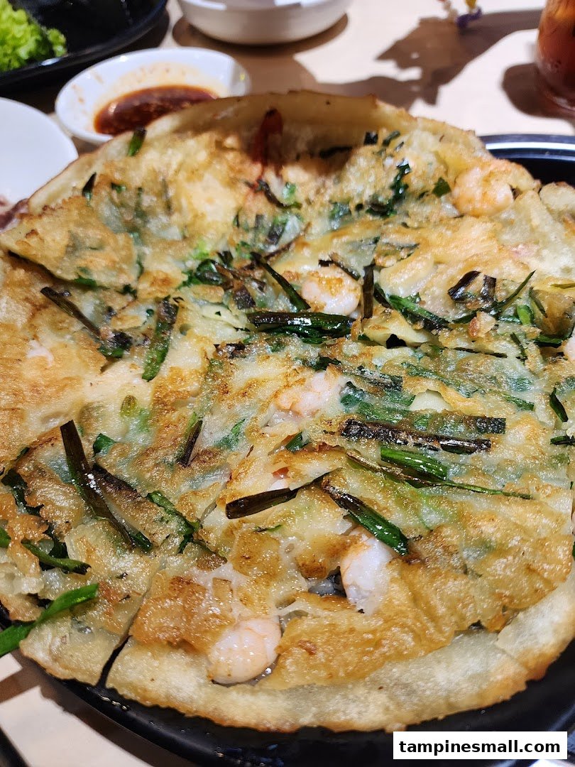 A close-up of a golden-brown Korean seafood and green onion pancake (haemul pajeon) on a black plate.