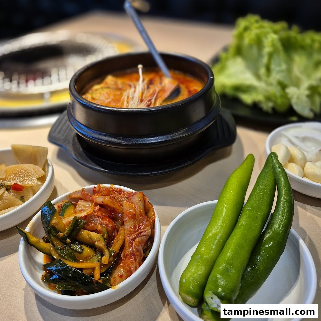 A close-up of a delicious Korean stew with an array of fresh banchan, including kimchi and green peppers.