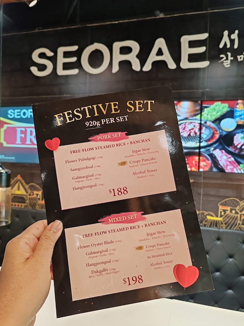 A hand holds a menu displaying 'Festive Set' options for Korean BBQ, with the 'Seorae' sign in the background.