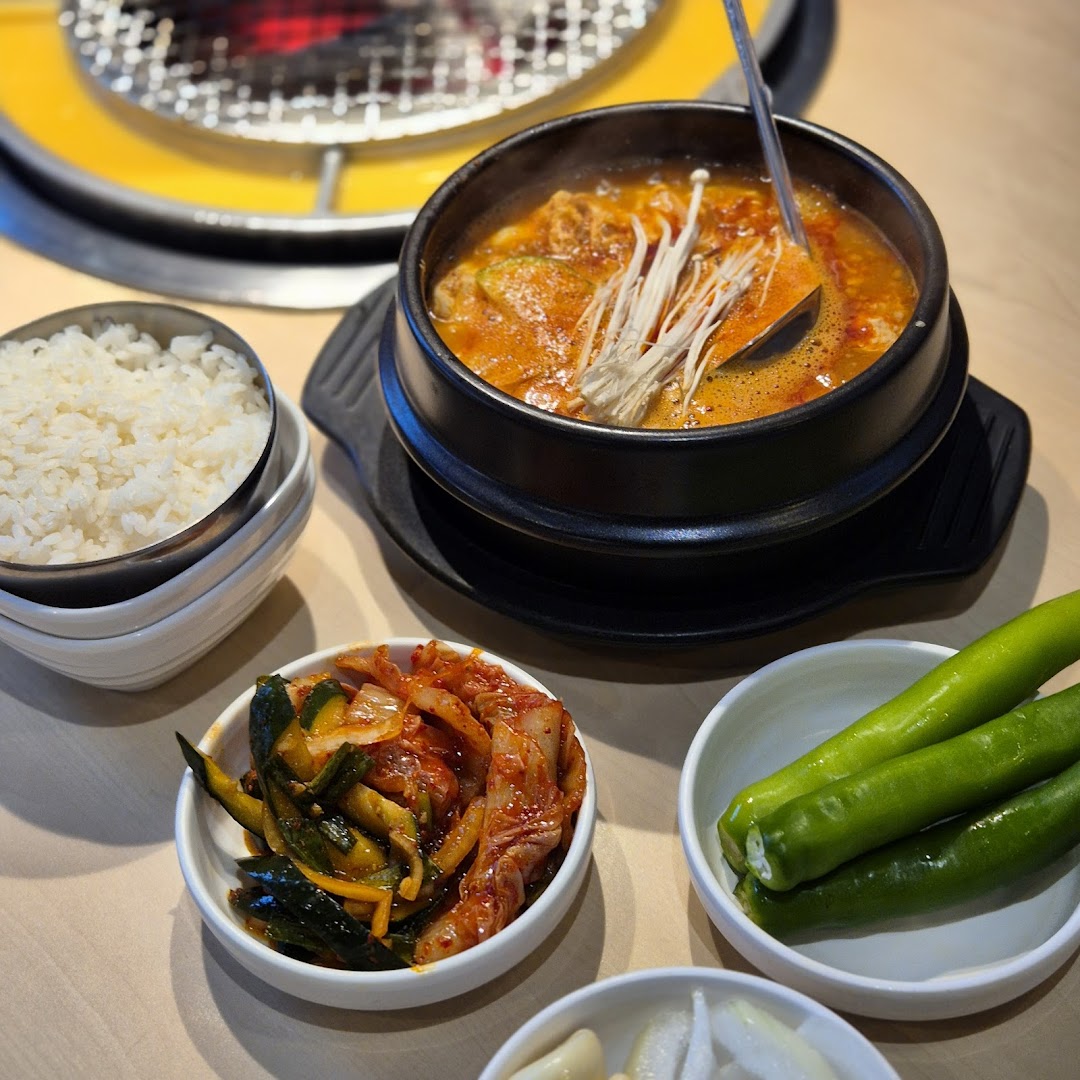 A steaming Korean stew with rice and banchan, set on a table with a grill in the background.