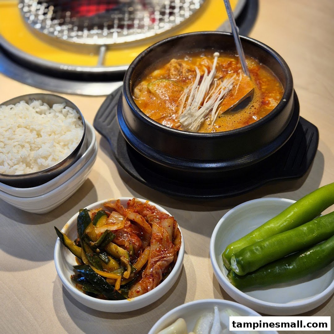 A steaming Korean stew with rice and banchan, set on a table with a grill in the background.