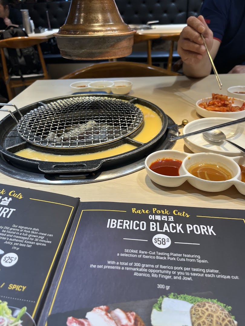 A Korean BBQ table with a grill, side dishes, and an open menu displaying premium pork cuts.