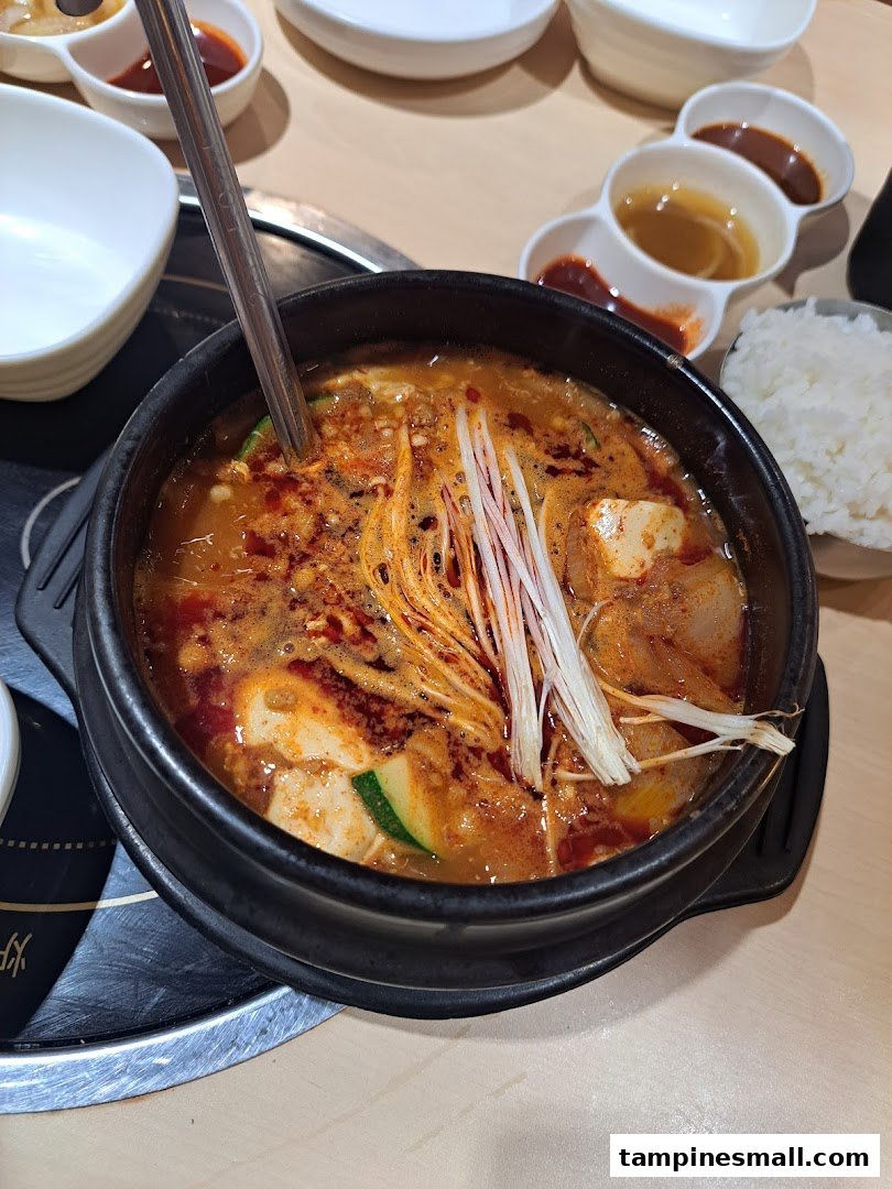 A close-up of a bubbling Korean hot pot with tofu, vegetables, and mushrooms, served with rice and side dishes.