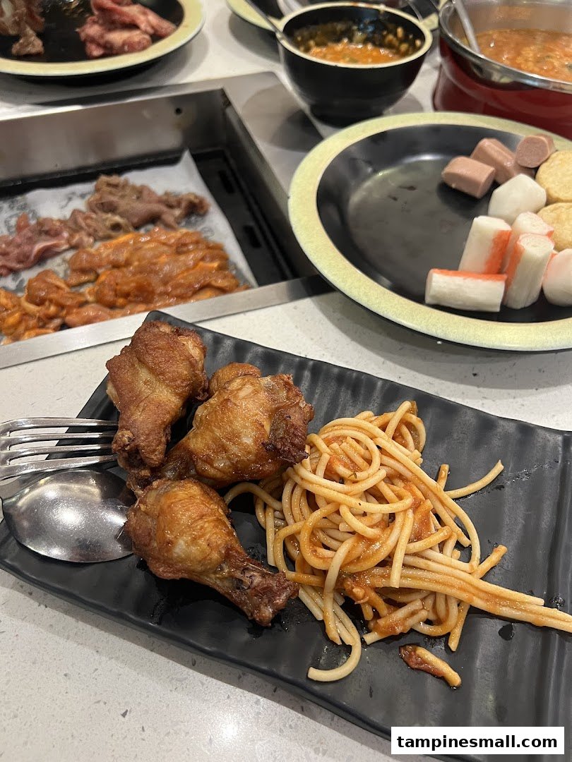 A close-up of fried chicken wings and spaghetti on a plate, with hotpot ingredients and raw meats in the background.