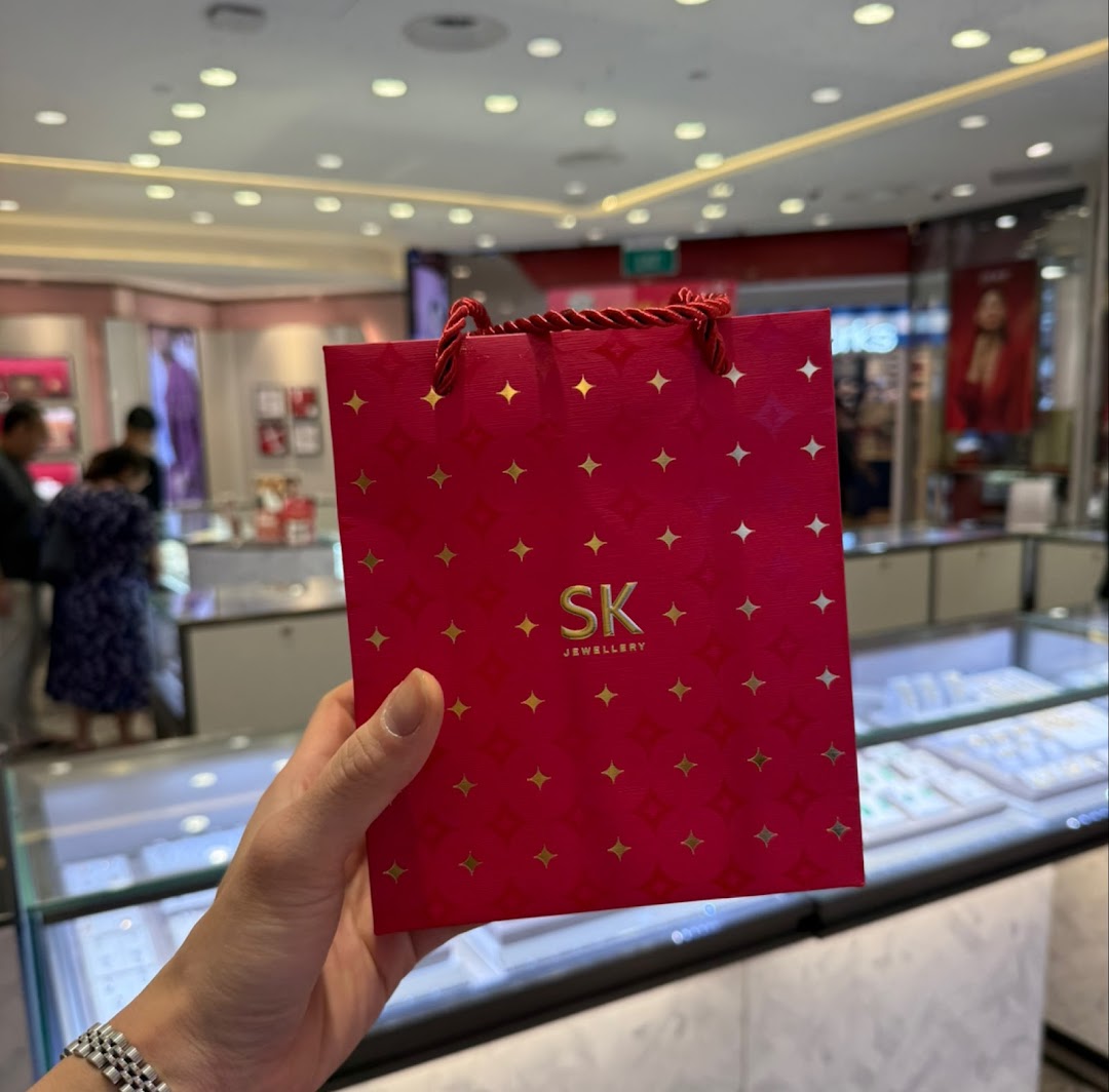 A hand holds a red SK Jewellery shopping bag inside a brightly lit jewellery store.