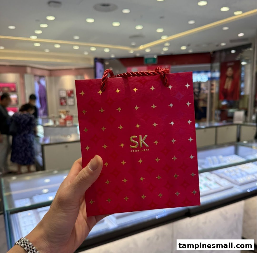 A hand holds a red SK Jewellery shopping bag inside a brightly lit jewellery store.