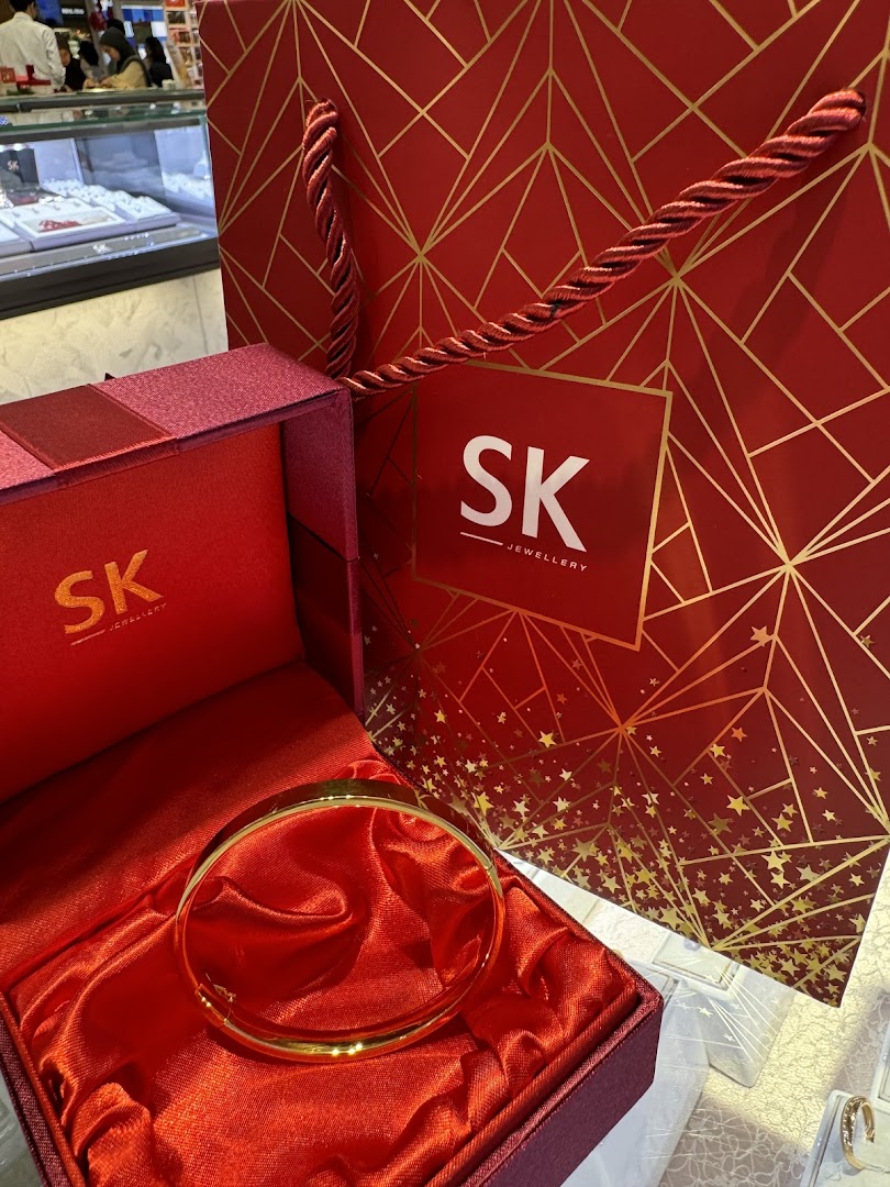 A gold bangle displayed in a red satin-lined gift box, with an SK Jewellery shopping bag.