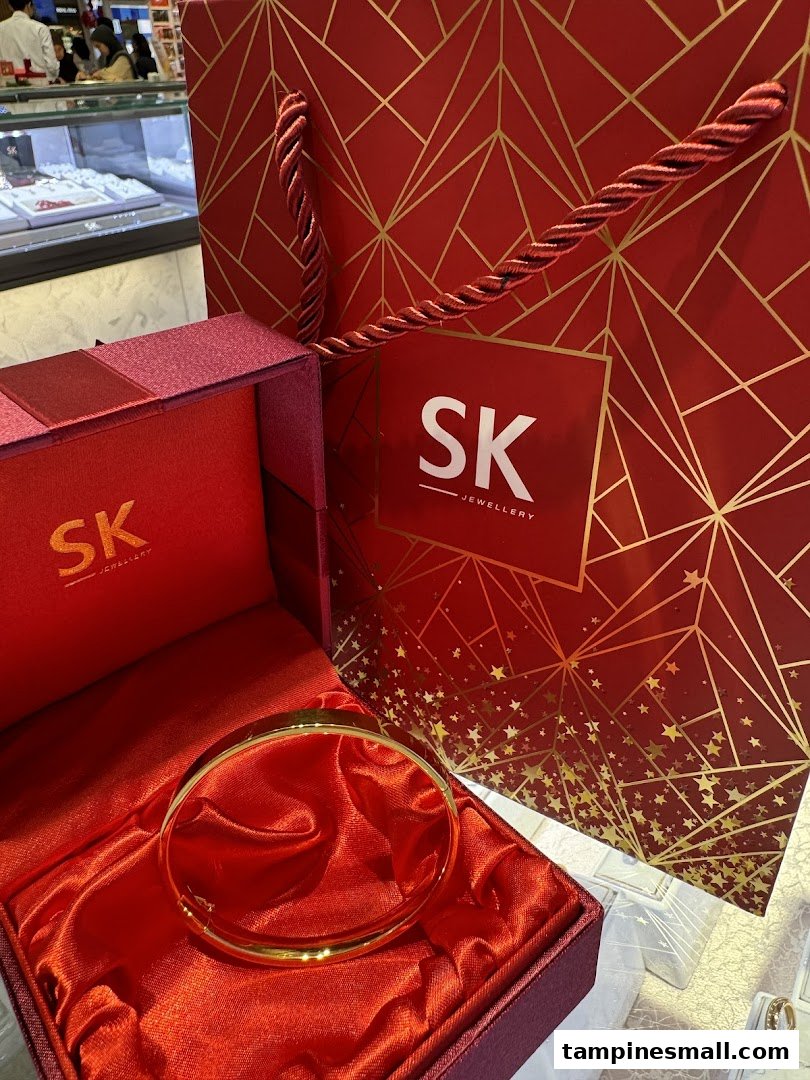 A gold bangle displayed in a red satin-lined gift box, with an SK Jewellery shopping bag.