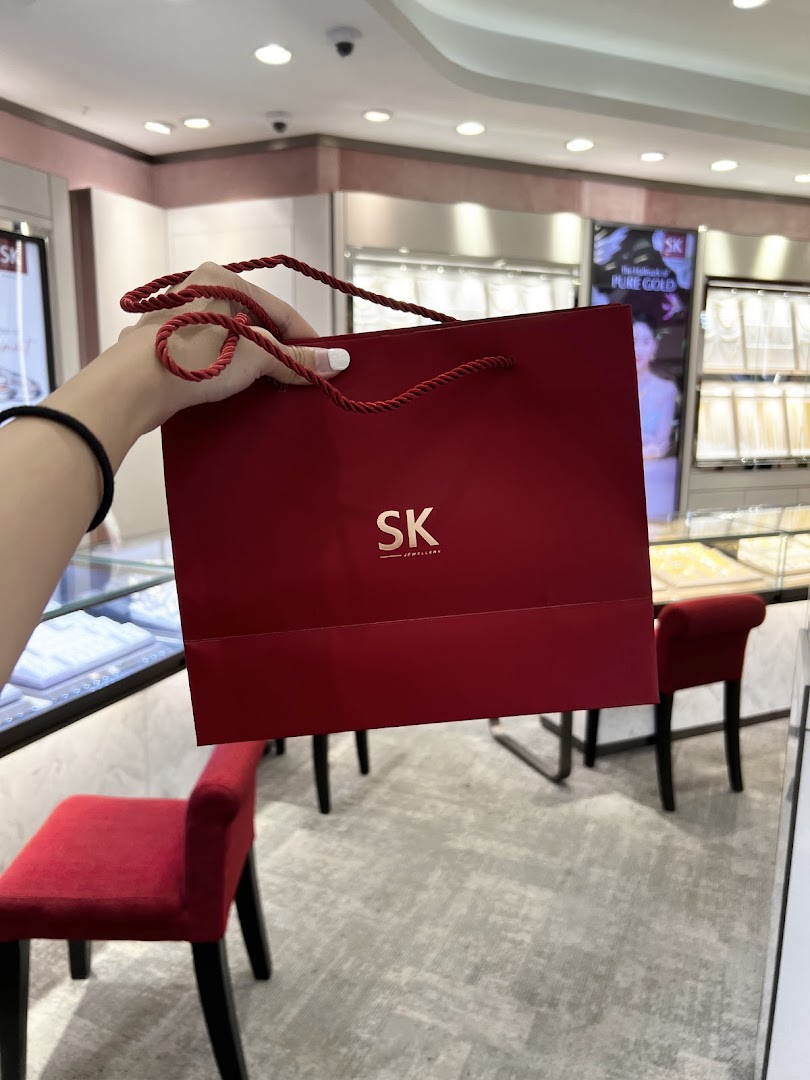 A person holds a red SK Jewellery shopping bag inside the store, showcasing the brand's packaging.