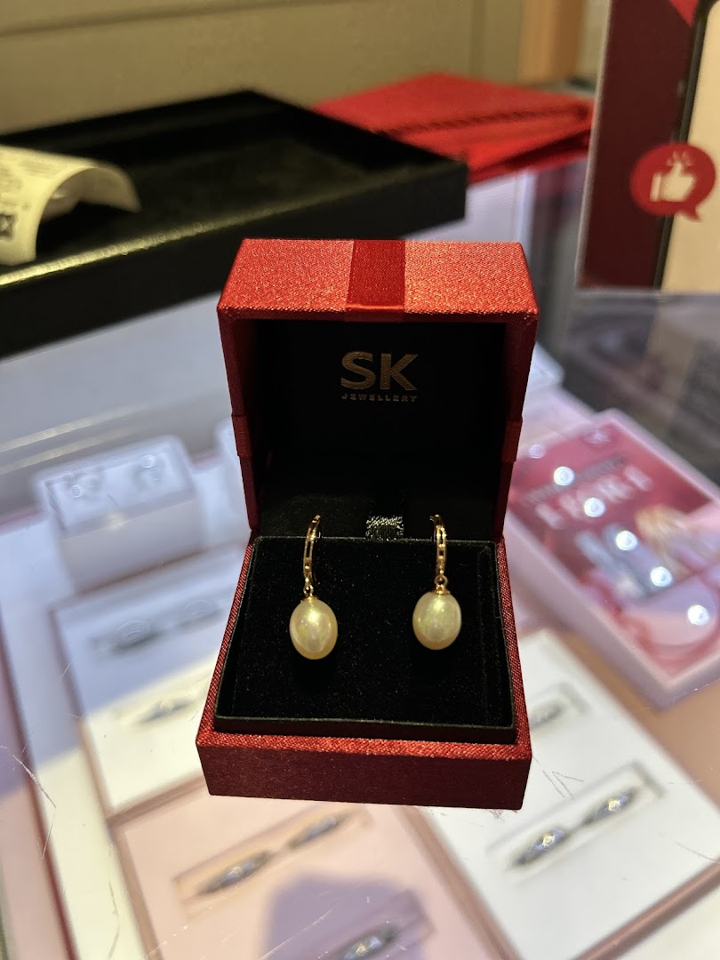 A pair of elegant pearl earrings displayed in a red SK Jewellery box on a glass counter.