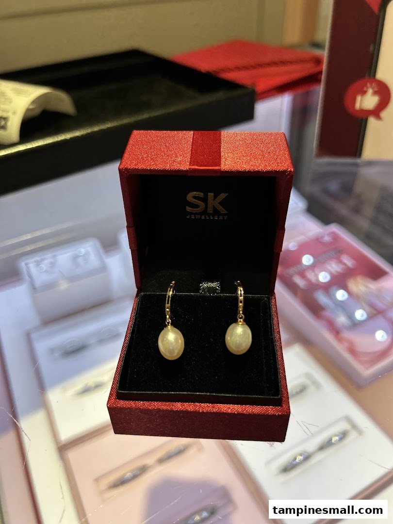A pair of elegant pearl earrings displayed in a red SK Jewellery box on a glass counter.