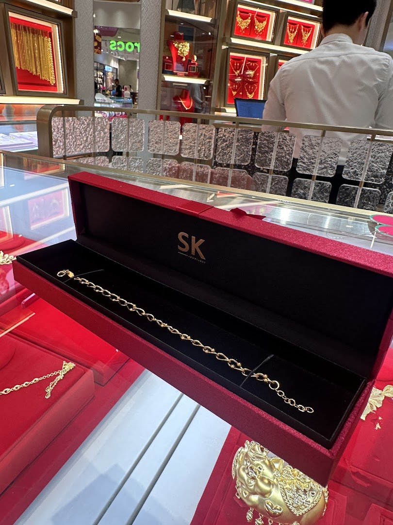 A gold bracelet displayed in a red SK Jewellery box amidst other gold ornaments in a brightly lit store.