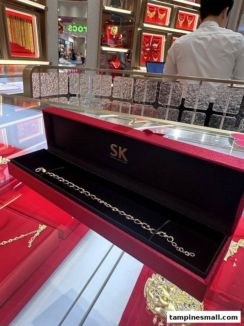A gold bracelet displayed in a red SK Jewellery box amidst other gold ornaments in a brightly lit store.